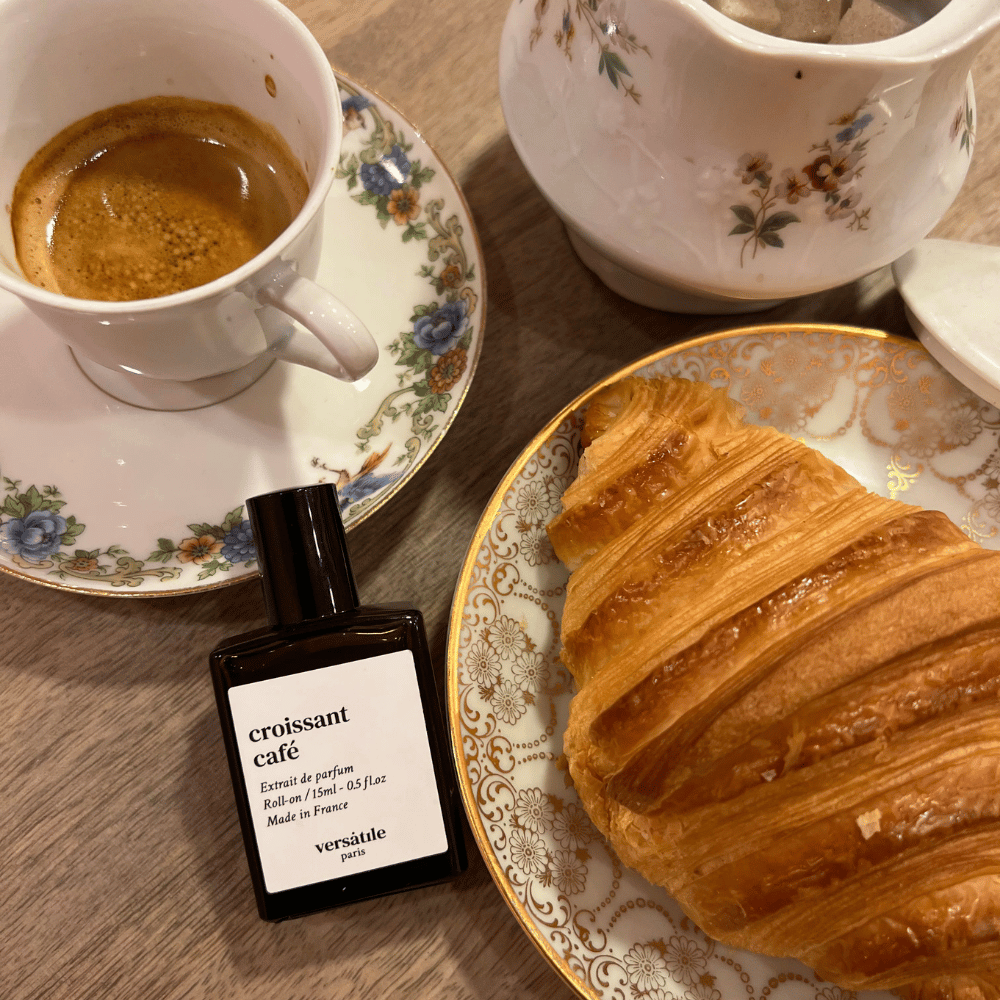 Au centre le flacon de parfum croissant café de la marque Versatile entouré des éléments qui composent le parfum : croissant, tasse de café pour une mise en situation