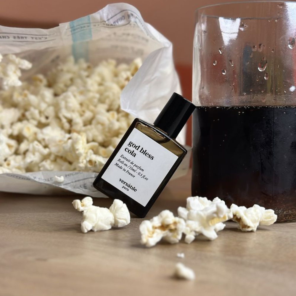 Le flacon de God Bless Cola de la marque Versatile Paris au centre posé contre un verre de cola. Dans le fond, un sachet de popcorn au caramel.