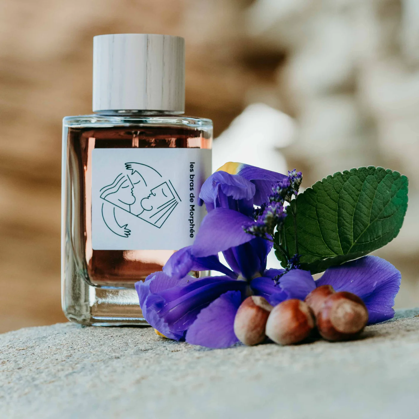 Au centre le flacon d'eau de parfum Les Bras de Morphée de la marque Hellenist entouré des éléments qui le composent : violette, noisette et iris