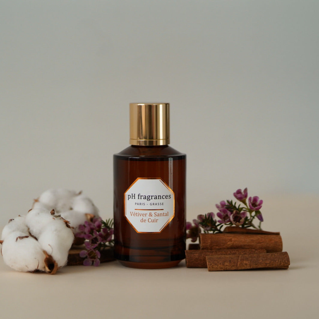 Au centre le flacon de 100ml d'eau de parfum Vetiver & Santal de Cuir de la marque pH Fragrances mis en scène avec les éléments qui le composent : santal, cuir et vétiver