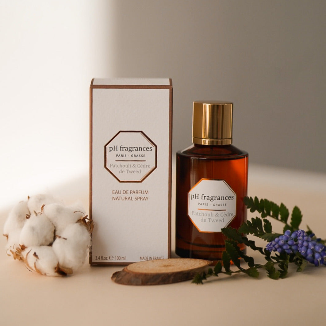 Au centre le flacon de 100ml d'eau de parfum Patchouli & Cèdre de Tweed de la marque pH Fragrances mis en scène avec les éléments qui le composent : patchouli et bois de cèdre